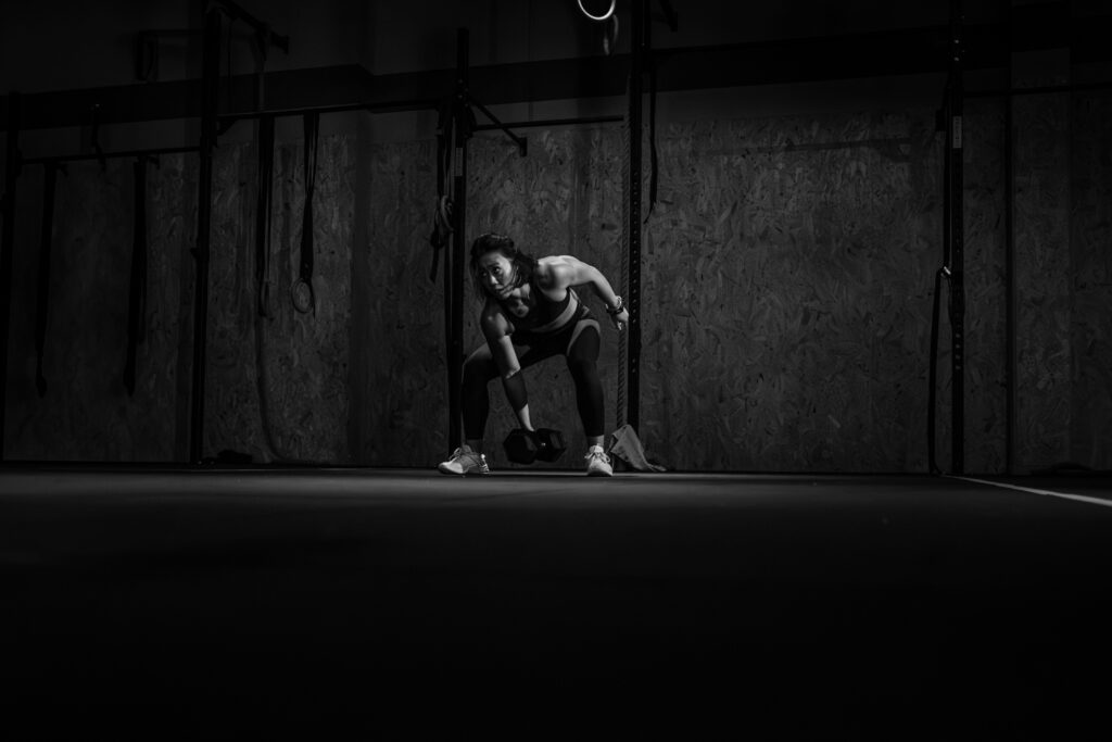 Une femme athlétique s'apprête à soulever un haltère, son visage concentré et ses muscles contractés, dans une salle de sport sombre et éclairée ponctuellement, le tout en noir et blanc.