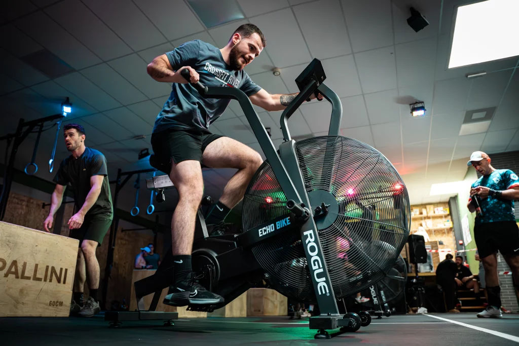 Personne en train de s’entraîner sur un air bike dans une salle de sport, avec d’autres athlètes autour.