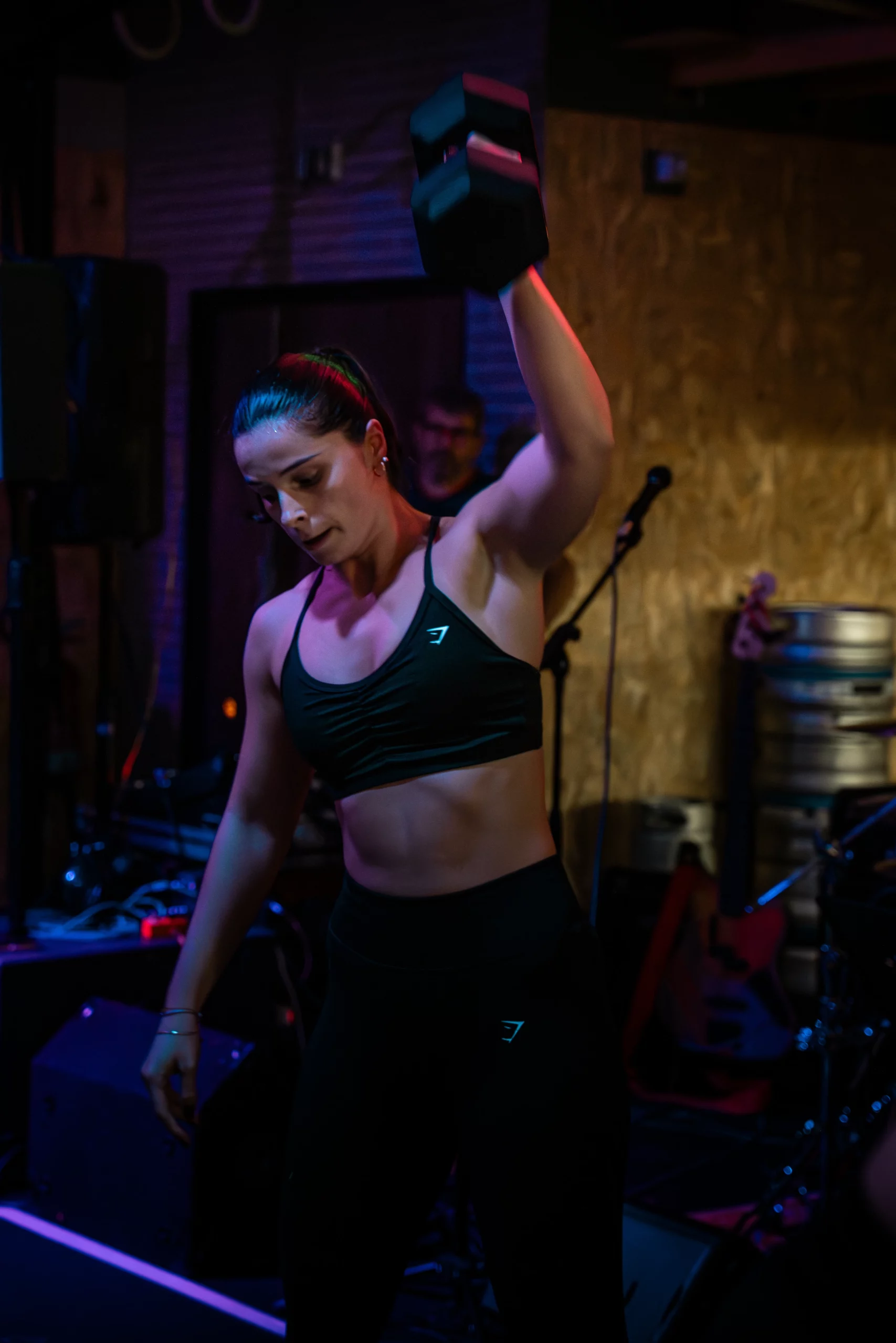 Une femme, vêtue d'un soutien-gorge de sport et d'un legging noirs, est photographiée en pleine action, levant un haltère au-dessus de sa tête avec son bras droit. Son expression est concentrée et on aperçoit de la sueur sur son front. La scène se déroule dans un lieu faiblement éclairé par des lumières colorées (tons violets et bleus), créant une ambiance dynamique, avec du matériel de scène (enceintes, micro) visible en arrière-plan.