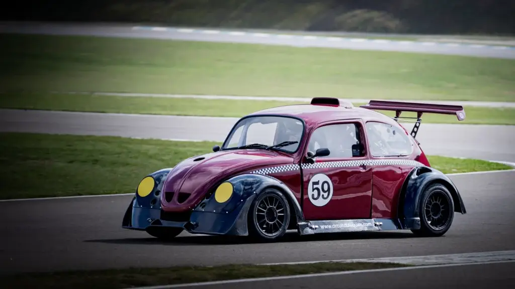 Résultat de la description de l'image L'image présente une Coccinelle Volkswagen modifiée pour la course sur une piste. La voiture est peinte en bordeaux et présente un damier noir et blanc le long de ses côtés. Le numéro "59" est bien en évidence sur la portière, et elle est équipée d'un aileron arrière proéminent. Le véhicule est également doté d'un kit de carrosserie personnalisé, avec des ailes avant et arrière plus larges, ainsi qu'un aileron avant. Les phares sont recouverts de film jaune. La voiture est photographiée en plein mouvement sur une piste de course bordée d'herbe.